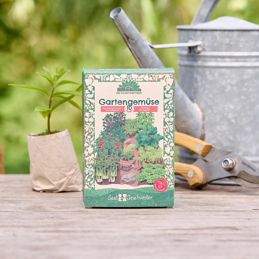 Saatgut-Set Gartengemüse von Die Stadtgärtner – Verpackung, im Hintergrund eine Gießkanne und Gartenschere