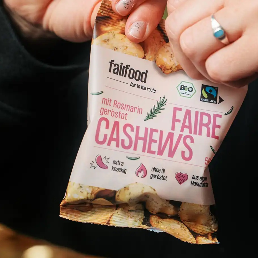 Person reißt fairfood – Cashews mit Rosmarin 50 g Tütchen auf
