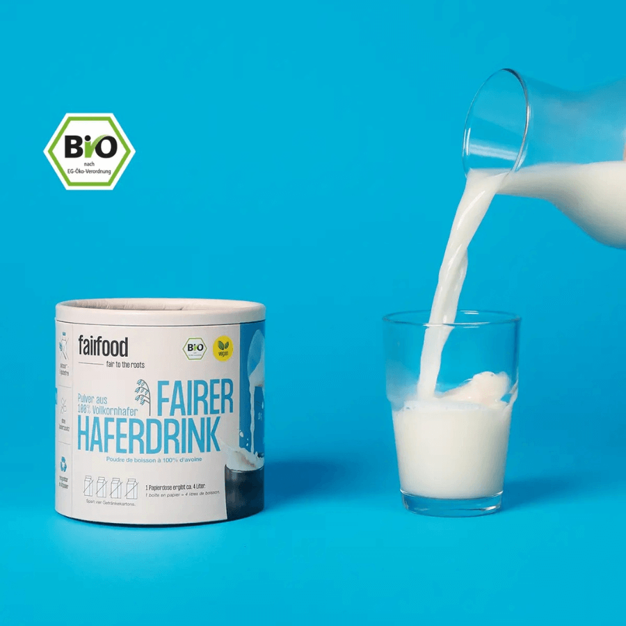 fairfood Haferdrinkpulver in der Dose, daneben wird aus einer Karaffe Milch in ein Glas gegossen