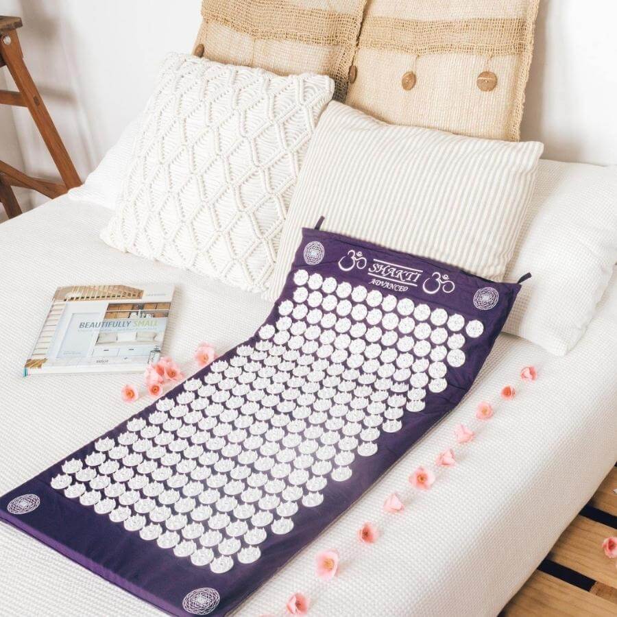 ShaktiMat Akupressurmatte Advanced auf einem Bett, umgeben von Kissen und Rosenblättern für Entspannung.