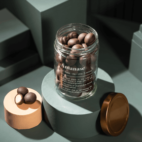 Amanase Bio-Macadamia im geöffneten Glas, links zwei Macadamia-Hälften und eine ganze