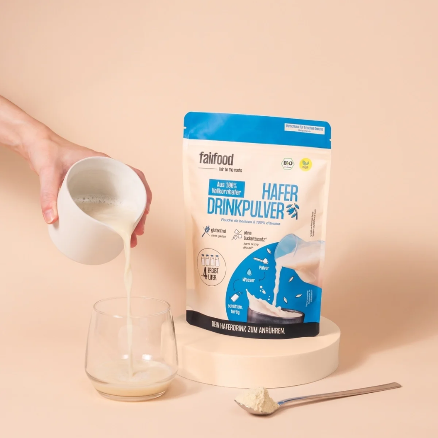 Person gießt sich Hafermilch in ein Glas, im Hintergrund steht der fairfood Haferdrinkpulver im 360g Beutel