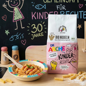 Bio Fusilli von social product – Verpackung steht auf einem Tisch neben einem Teller gekochter Nudeln