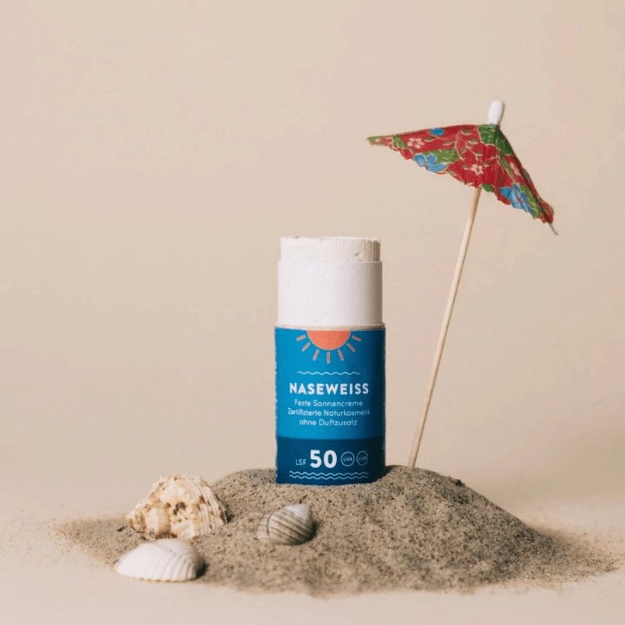 4peoplewhocare feste Sonnencreme LSF 50 Verpackung auf Sandhäufchen mit Muscheln und mit Schirm 